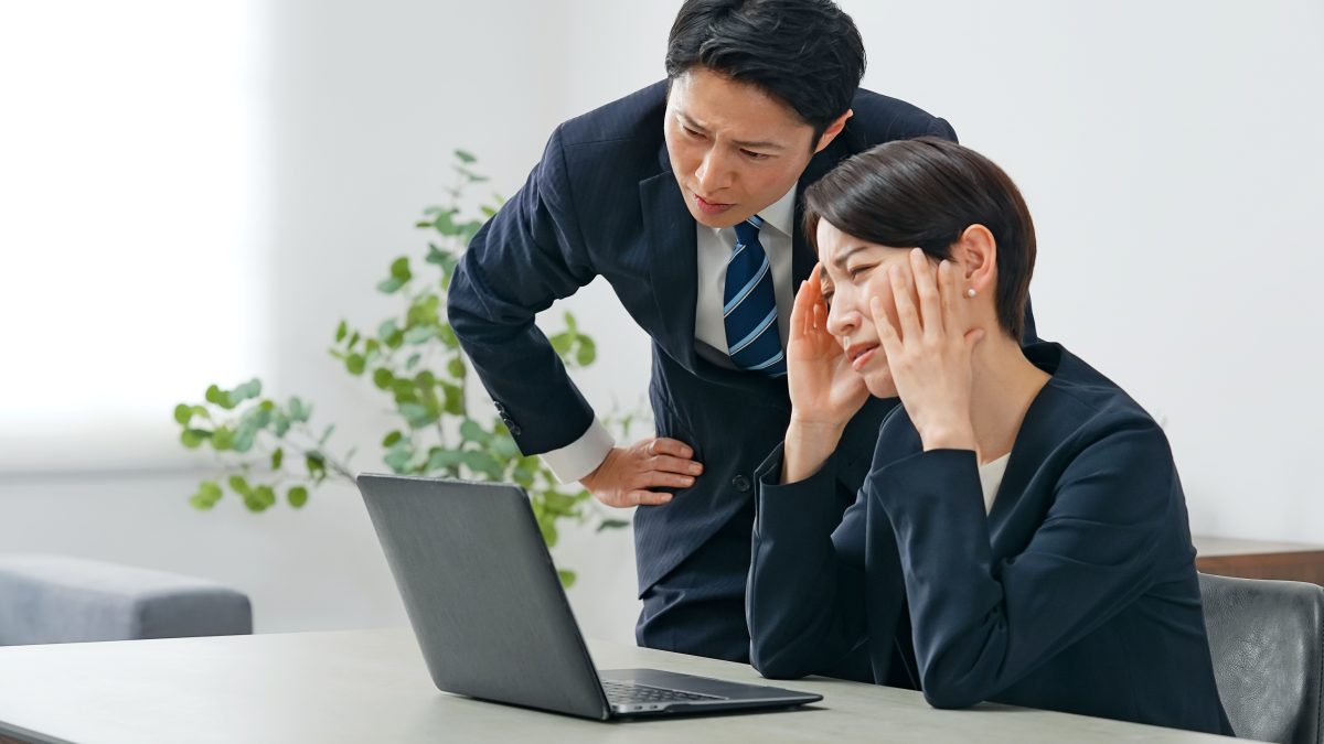 オフィスらしき空間で、ノートパソコンを見つめる女性が両手で頭を抱え、困惑またはストレスを感じている様子です。その横にはスーツ姿の男性が心配そうに、あるいは厳しく、ノートパソコンの画面を覗き込んでいます。これは、職場の問題、システム障害、緊急の課題、またはビジネス上の困難な状況を共有している場面を示唆しています。