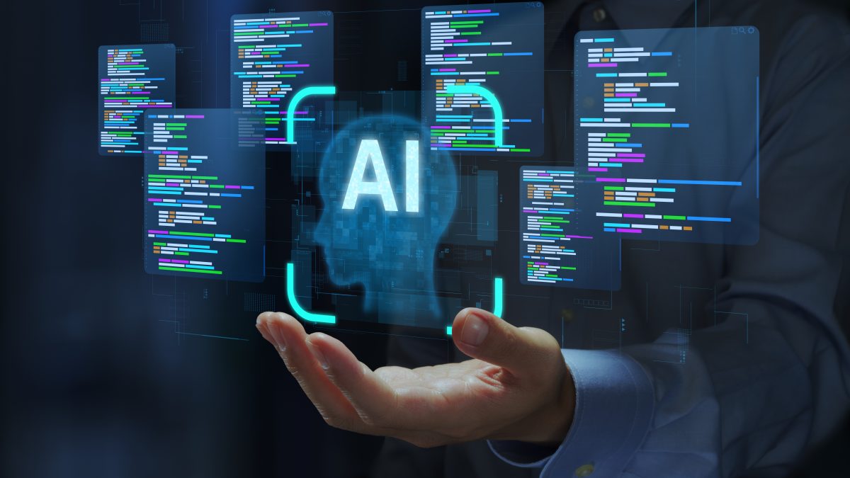 スーツを着用した人物が手を差し出し、その手のひらの上に、青い光で「AI」の文字と脳のネットワークを模した人間の頭部のシルエットが浮かび上がっています。その周囲には、プログラミングコードが表示された複数の半透明なウィンドウが浮遊しており、人工知能（AI）の開発、機械学習のコーディング、または未来のテクノロジーとイノベーションの概念を示唆しています。