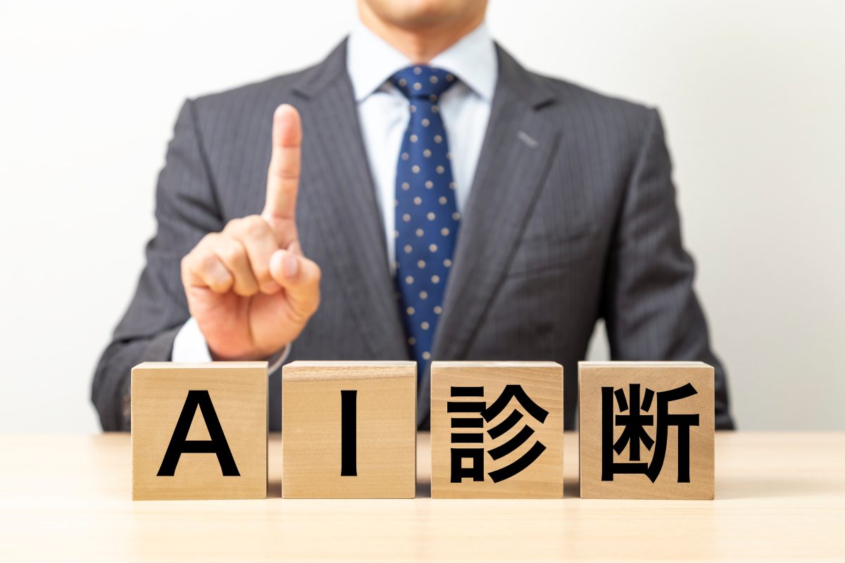 ビジネススーツを着用した人物が、指を立てて何かを指示しています。手前の木製テーブル上には、「AI」と漢字の「診断」の文字がそれぞれ刻まれた4つの木製ブロックが並んでおり、人工知能（AI）による診断サービス、コンサルティング、または専門的な技術提案の概念を表現しています。